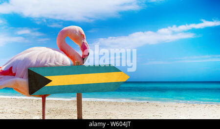 Bahamas Flagge auf hölzernen Tisch am Strand Hintergrund mit rosa Flamingo. Es liegt am Strand und klares Wasser von Meer und blauer Himmel im Hintergrund. Es ist Stockfoto