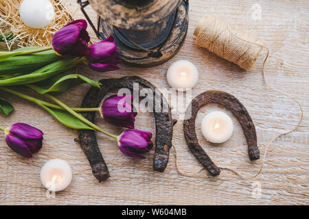 Flach auf zwei rostige Kleine und Große Hufeisen auf Holzbrett Hintergrund, mit kleinen weißen Kerzen und lila Tulpen eingerichtet, Feder Stockfoto