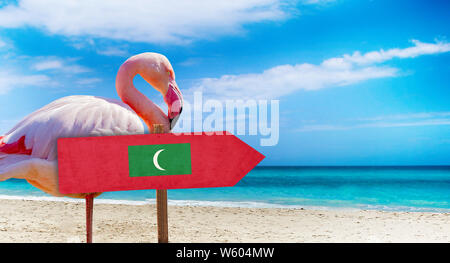 Malediven Flagge auf hölzernen Tisch am Strand Hintergrund mit rosa Flamingo. Es liegt am Strand und klares Wasser von Meer und blauer Himmel im Hintergrund. Es ist Stockfoto