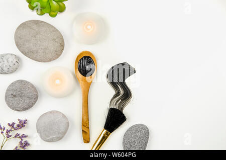 Flach auf Schlamm Holzkohle Maske auf Holzlöffel und Abstrich auf weißem Hintergrund, mit Schönheit Pinsel und flache Meer Steine und spa Kerzen umgeben. Ro Stockfoto