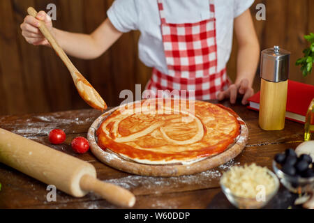 Mädchen tragen karierte Schürze und Kopfbügel kochen Pizza durch die Verbreitung der Sauce mit Löffel in Herz Form auf Tabelle mit Zutaten für Pizza, in stilvollen Küche aus Holz. Stockfoto