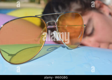 Junge Frau schlafend 7 Sonnenbaden auf eine lilo in einem Schwimmbad. Metall gelb Sonnenbrille im Vordergrund eingerahmt. Sommer vibes, Freizeit, Spaß und Freizeit Stockfoto