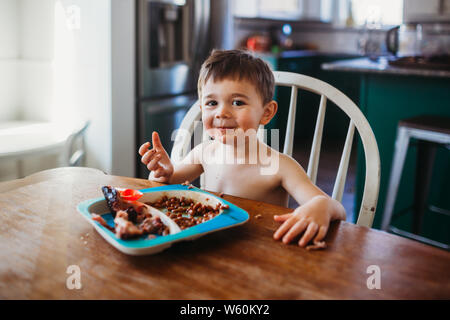 Junge lächelnd mit Essen am Esstisch Stockfoto