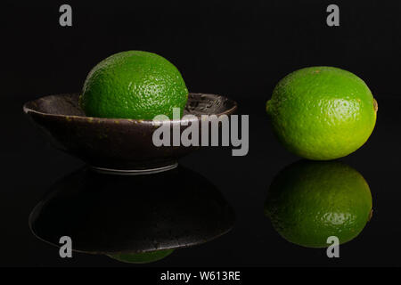 Gruppe von zwei ganze Frische grüne Kalk ist man beiseite und der andere ist in einem dunklen Keramik Schüssel auf schwarzem Glas isoliert Stockfoto