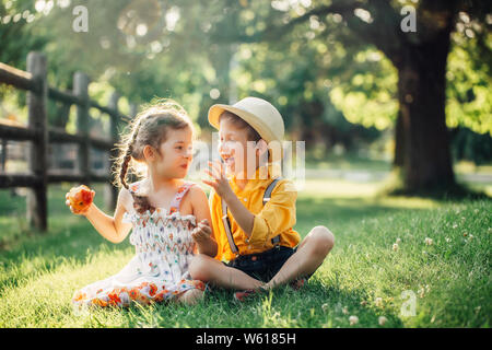 Kaukasische Kinder Jungen und Mädchen Geschwister zusammen sitzen, die gemeinsame Nutzung von Apple. Zwei Kinder Bruder und Schwester Essen süße Frucht im Park im Sommer Tag. Beste fr Stockfoto