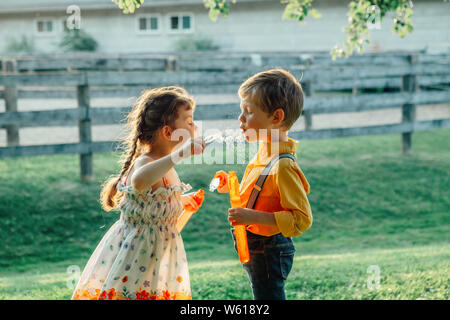 Lustige kaukasischen Kinder Mädchen und Jungen bläst Seifenblasen im Park im Sommer Sonnenuntergang. Authentische glückliche Kindheit Moment der Freundschaft. Lifestyle ac Stockfoto