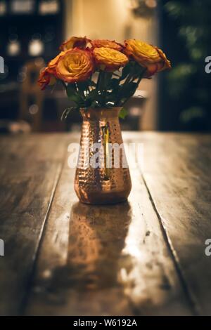 Nahaufnahme von orangen Rosen in einer kleinen Metallvase Auf einem runden Holztisch mit verschwommenem Hintergrund Stockfoto
