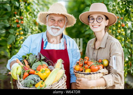 Porträt einer jungen Frau mit Großvater holding Körbe voll mit frischem Gemüse Ernte im Gewächshaus eines kleinen landwirtschaftlichen Betrieb Stockfoto