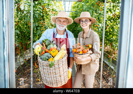 Porträt einer jungen Frau mit Großvater holding Körbe voll mit frischem Gemüse Ernte im Gewächshaus eines kleinen landwirtschaftlichen Betrieb Stockfoto