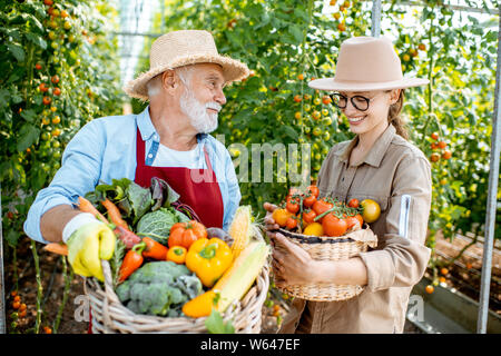 Porträt einer jungen Frau mit Großvater holding Körbe voll mit frischem Gemüse Ernte im Gewächshaus eines kleinen landwirtschaftlichen Betrieb Stockfoto