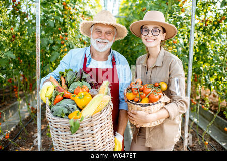 Porträt einer jungen Frau mit Großvater holding Körbe voll mit frischem Gemüse Ernte im Gewächshaus eines kleinen landwirtschaftlichen Betrieb Stockfoto
