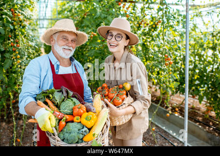 Porträt einer jungen Frau mit Großvater holding Körbe voll mit frischem Gemüse Ernte im Gewächshaus eines kleinen landwirtschaftlichen Betrieb Stockfoto