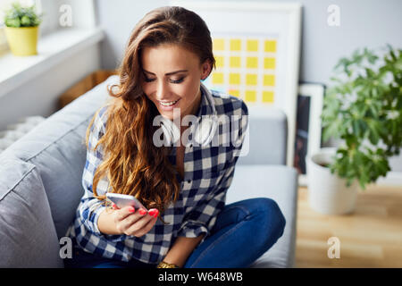 Freundliche hübsche junge Frau auf dem Sofa zu Hause sitzen, lachen und über Telefon Stockfoto