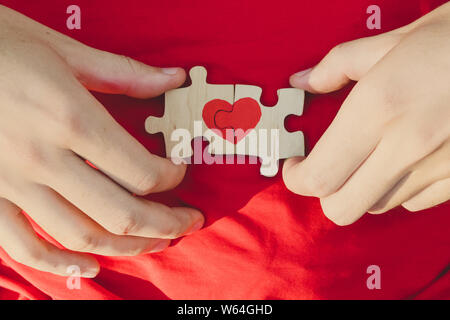 Rotes Herz ist auf die Teile des Puzzles in männlichen Händen auf rotem Hintergrund dargestellt. Liebe Konzept. St. Valentinstag Stockfoto