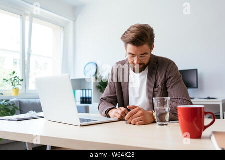 Mann mit Smartphone, während am Schreibtisch sitzt mit Laptop im Büro zu Hause Stockfoto
