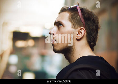 Perky findiger Kerl in der Sonnenbrille und schwarze Kleidung, schaut auf, offensichtlich etwas denken, und leckt mit seiner Zunge die Lippen. Stockfoto