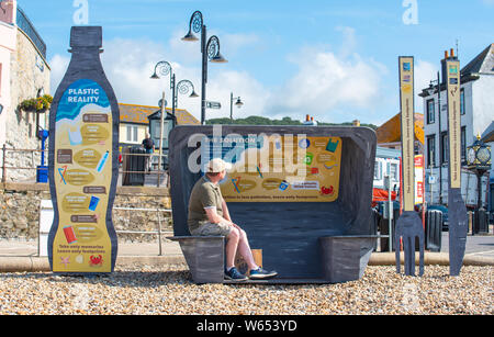 Lyme Regis, Dorset, Großbritannien. 31. Juli 2019. UK Wetter: ein Mann bewundert die Aussicht aus einem riesigen Wurf Skulptur von Takeaway Verpackung hat hat am malerischen Strand errichtet worden in den Badeort Lyme Regis. Bestehend aus 8 Fuß hoch Kunststoff Trinkflasche, Besteck 6 Fuß hoch und einem Chip/burger Box 4 x 4 Fuß die Skulpturen die Bemühungen der Wurf frei Küste und Meer und Partner bei der Förderung ökologisch verantwortungsvolle Entscheidungen zu fördern. Es ist zu hoffen, dass die vorübergehende Installation von architektonischen Designer Jack Hebeny ermutigen wird, die Besucher ihren Müll mit nach Hause zu nehmen. Gutschrift: Stockfoto