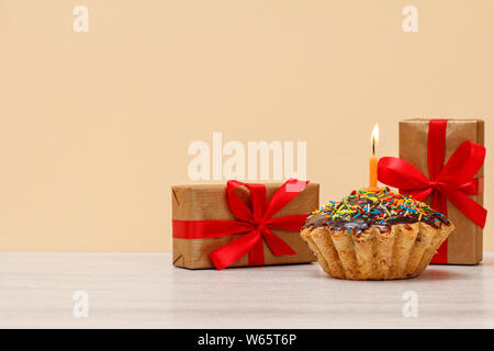 Lecker geburtstag Muffin mit Schokoladenüberzug und Karamell, mit brennenden festliche Kerze und Geschenkboxen mit roten Bändern auf dem beigen Hintergrund eingerichtet. Stockfoto