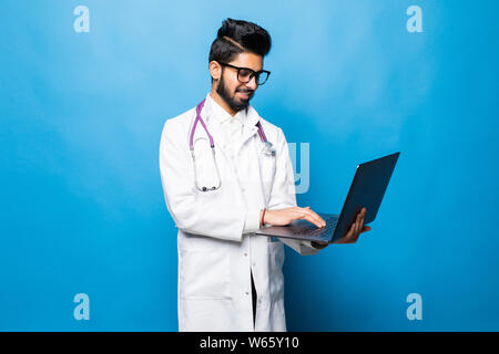 Indische Arzt, der im Studio während der Arbeit mit dem Laptop, auf blauem Hintergrund isoliert Stockfoto