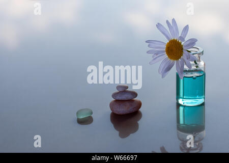 Eine Pyramide von Steinen und eine Flasche Kamille auf einer reflektierenden Oberfläche stecken. Es ist eine blaue Flüssigkeit in der Flasche. Selektiver Fokus auf die Blume. Ther Stockfoto