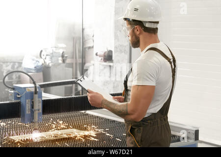 Ansicht von der Seite des männlichen Ingenieur tragen Uniform, Schutzbrille und Helm, Ordner und Plasmaschneiden. Muskulöse mann Kontrolle von Geräten und Anlagen. Stockfoto