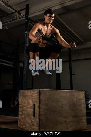 Eine athletische kurze behaarte Frau mit starken abs tut, springt in ein Fitnessstudio. Die weiblichen Athleten lächelt und springen auf die Box. Schwarz und Weiß. Stockfoto