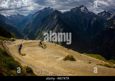 LES DEUX ALPES, Frankreich. Zwei Mountainbiker, eine 'bermed' Ecke auf einem Bike-park Trail, mit einem steilen Flußtal und dramatische Alpengipfel hinter sich. Stockfoto