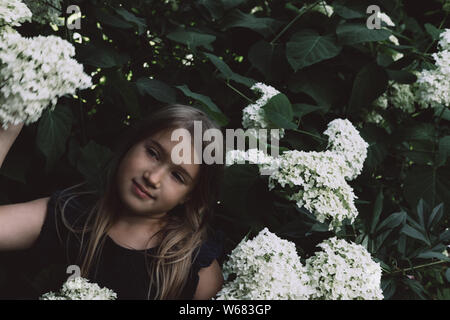 Süße kleine Mädchen in blühenden Hortensien. Person und Blumen auf dunklen natürlichen Hintergrund. Kind mit Blumenstrauß vor der Tür. Zu Park, Wald im Frühling und Sommer. Verbindung mit der Natur Idee Stockfoto