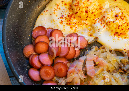 Rührei mit Speck, Zwiebeln und Würstchen. In der Pfanne auf den Tisch. Stockfoto