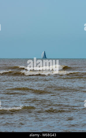 Ein Segelboot oder Yacht am Horizont mit Wellen am Ufer im Vordergrund. Stockfoto