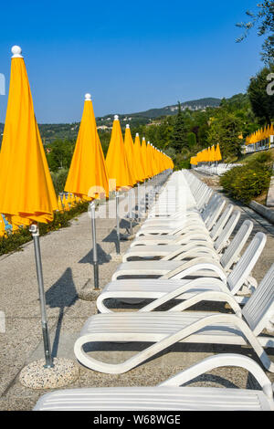 GARDA, Gardasee, Italien - September 2018: Lange Linie aus weißem Kunststoff Sonnenliegen und gelben Sonnenschirmen im Poiano Hotel & Spa in der Nähe des Gardasees. Stockfoto