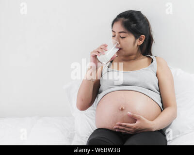 Asiatische schwangere Frau trinkt ein Glas Milch zu sitzen auf dem Bett zu Hause, Lifestyle Konzept. Stockfoto