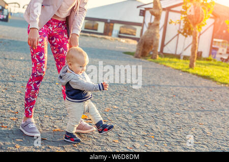 Süße kleine adorable Blonde toddler Boy machen erste Schritte mit Mutter Unterstützung im City Park am abend sonnenuntergang Zeit. Gerne lustig Kind laufen lernen Stockfoto