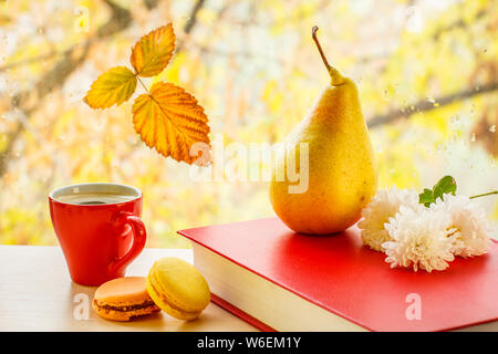 Tasse Kaffee mit Makronen, Birne und weißen Blumen auf dem Roten Buch und Fensterglas mit Wassertropfen in der verschwommenen natürlichen Hintergrund. Stockfoto