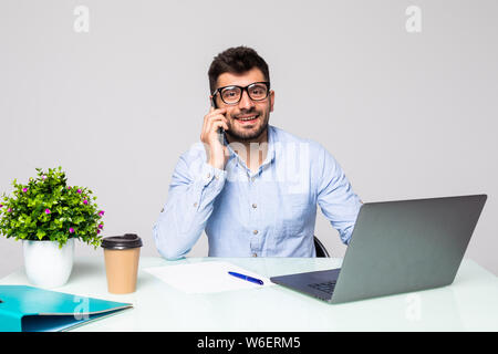 Gut aussehender Geschäftsmann in Brillen wird mithilfe eines Computers, auf dem Handy und lächelnd, während im Büro arbeiten Stockfoto