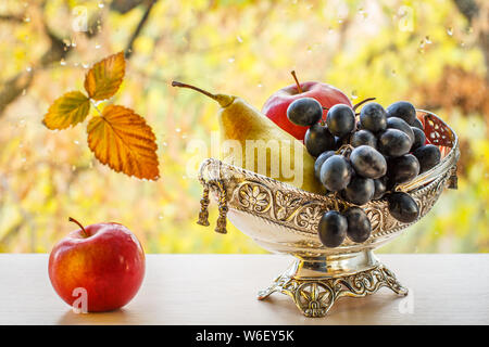 Apple und Metall Vase mit gelben Birnen und Trauben. Trockene Blatt auf Fensterglas mit Wassertropfen und Bäume im Herbst in den Hintergrund. Stockfoto