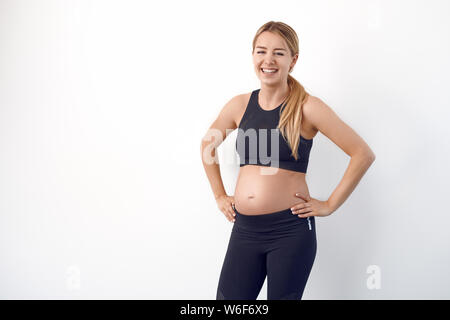 Glücklichen gesunden jungen schwangeren Frau in Schwarz Sportbekleidung auf den Hüften in die Kamera schaut mit einem strahlenden Lächeln Stockfoto