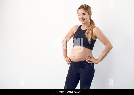 Glücklichen gesunden jungen schwangeren Frau in Schwarz Sportbekleidung auf den Hüften in die Kamera schaut mit einem strahlenden Lächeln Stockfoto