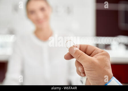 Nahaufnahme des goldenen Ehering, die Hand holding Mann mit unscharfen Hintergrund hinter im Schmuckladen. Älterer mann Kauf ihm neues Zubehör, dass kompetente weibliche Verkäufer empfohlen Stockfoto