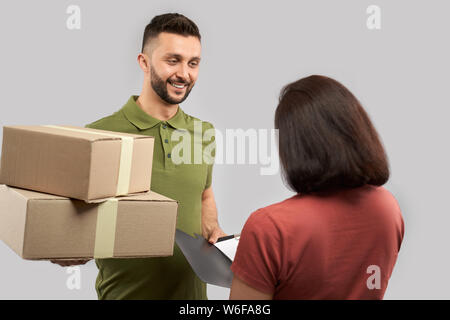 Jung, gutaussehend, lächelnd Delivery Man stehen und hält zwei Kartons und Ordner. Frau, Boxen und Anmeldung für den Eingang. Konzept der Lieferservice. Stockfoto