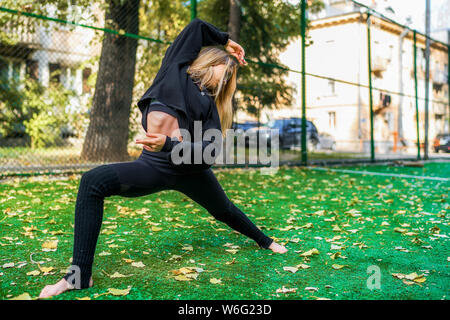 Junge blonde Frau in schwarzer Kleidung yoga Pose im Freien auf Fußballplatz in Georgien Stockfoto