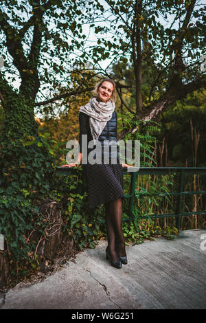 Hübsche junge Frau trägt ein Kleid und auf der Brücke von grünen Bäumen im Park umgeben, an der Kamera schaut lächelnd Stockfoto