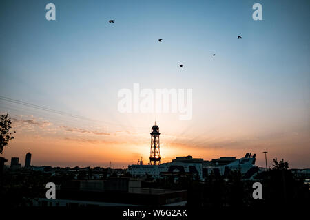 Montjuic Seilbahn Tower in der Dämmerung mit Sonne hinter dem Turm bei Sonnenaufgang mit spektakulären Strahlen, ein Wahrzeichen von Barcelona mit dem Hafen und der Kreuzfahrt sh Stockfoto