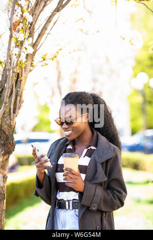 Charmante afro-amerikanische Frau mit Kaffee plaudern mit Freunden in sozialen Netzwerken und Surfen auf Internetseiten für die Suche nach interessanten Plätzen in Stockfoto