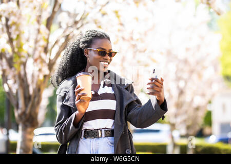 Charmante afro-amerikanische Frau mit Kaffee plaudern mit Freunden in sozialen Netzwerken und Surfen auf Internetseiten für die Suche nach interessanten Plätzen in Stockfoto
