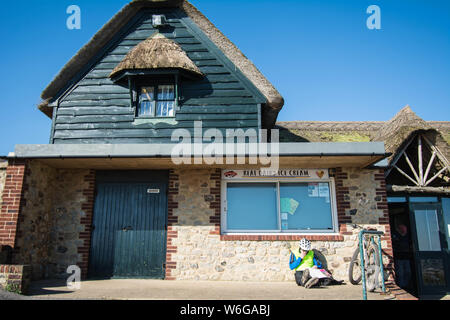 Branscombe Beach Shop, der Eiscreme verkauft, mit Radfahrer, der draußen auf der Treppe in Devon Fahrradhut Helmtür Holz grün bemalte Stein Hütte Stockfoto