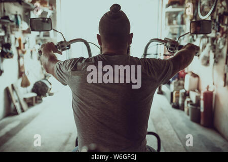 Schön brutalen Mann mit einem Bart sitzt auf einem Motorrad in seiner Garage, Ansicht von der Rückseite. Stockfoto
