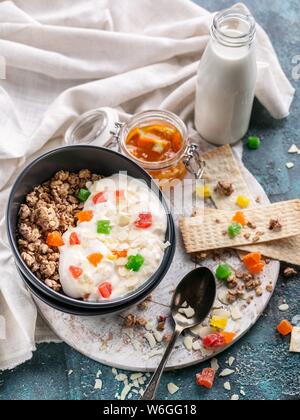 Müsli oder Müsli mit kandierten Früchten in einem dunklen Schüssel, hausgemachtes Joghurt, Orange Marmelade und Milch. Leckeres und gesundes Frühstück. Vertikaler Dampfstoß Stockfoto