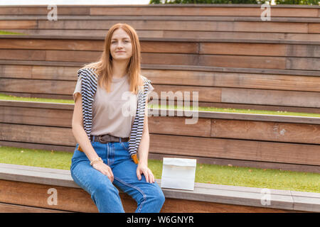 Lächelnde blonde Schüler Mädchen in T-Shirt und Blue Jeans sitzt auf einer Holzbank mit einer Doggy Bag auf einen sonnigen Tag im Park. Mittagspause. Leer Stockfoto