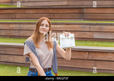 Lächelnde blonde Schüler Mädchen in T-Shirt und Blue Jeans sitzt auf einer Holzbank mit einer Doggy Bag auf einen sonnigen Tag im Park. Mittagspause. Leer Stockfoto